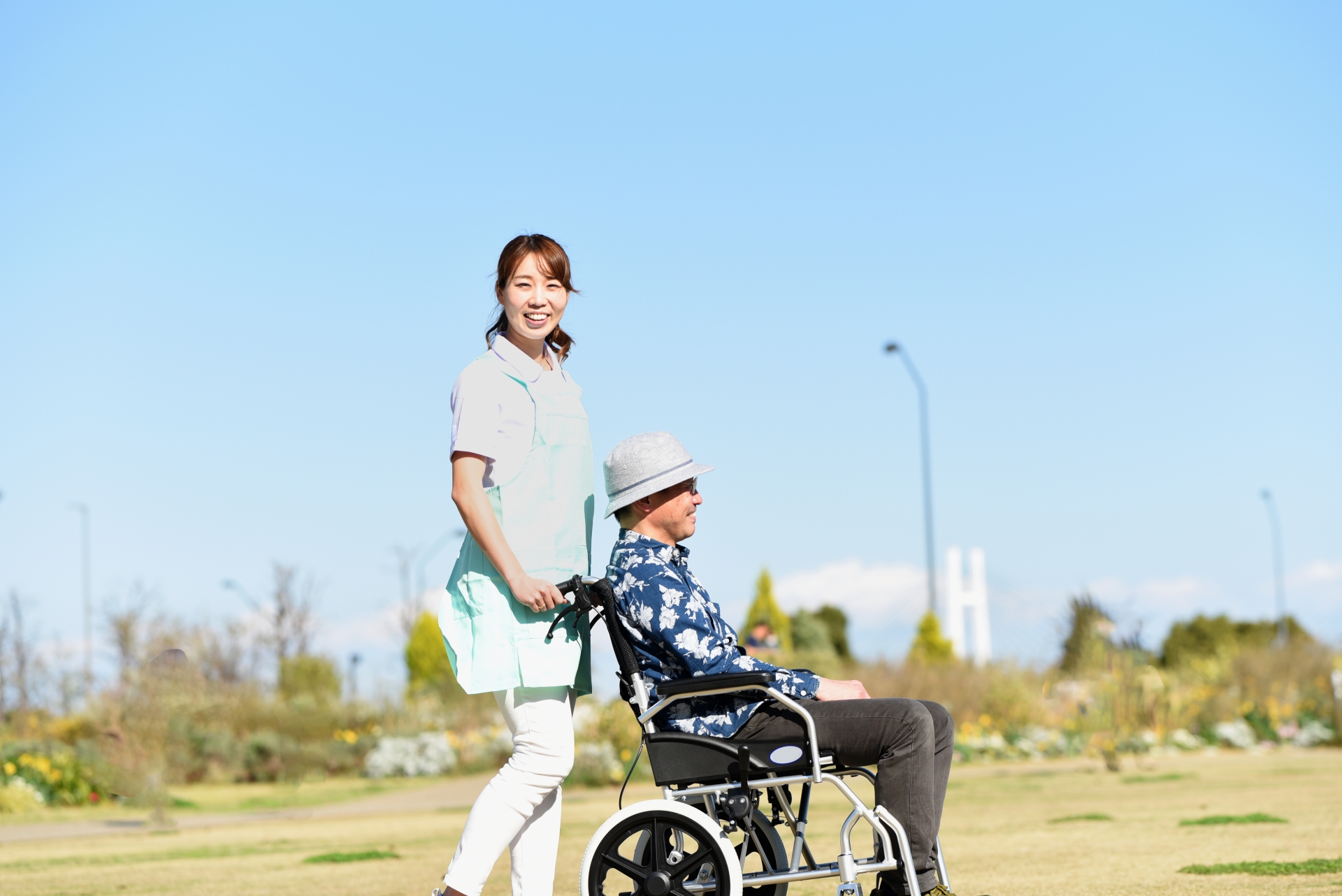 青空の屋外で車椅子を押す若い介護士の女性とシニア男性