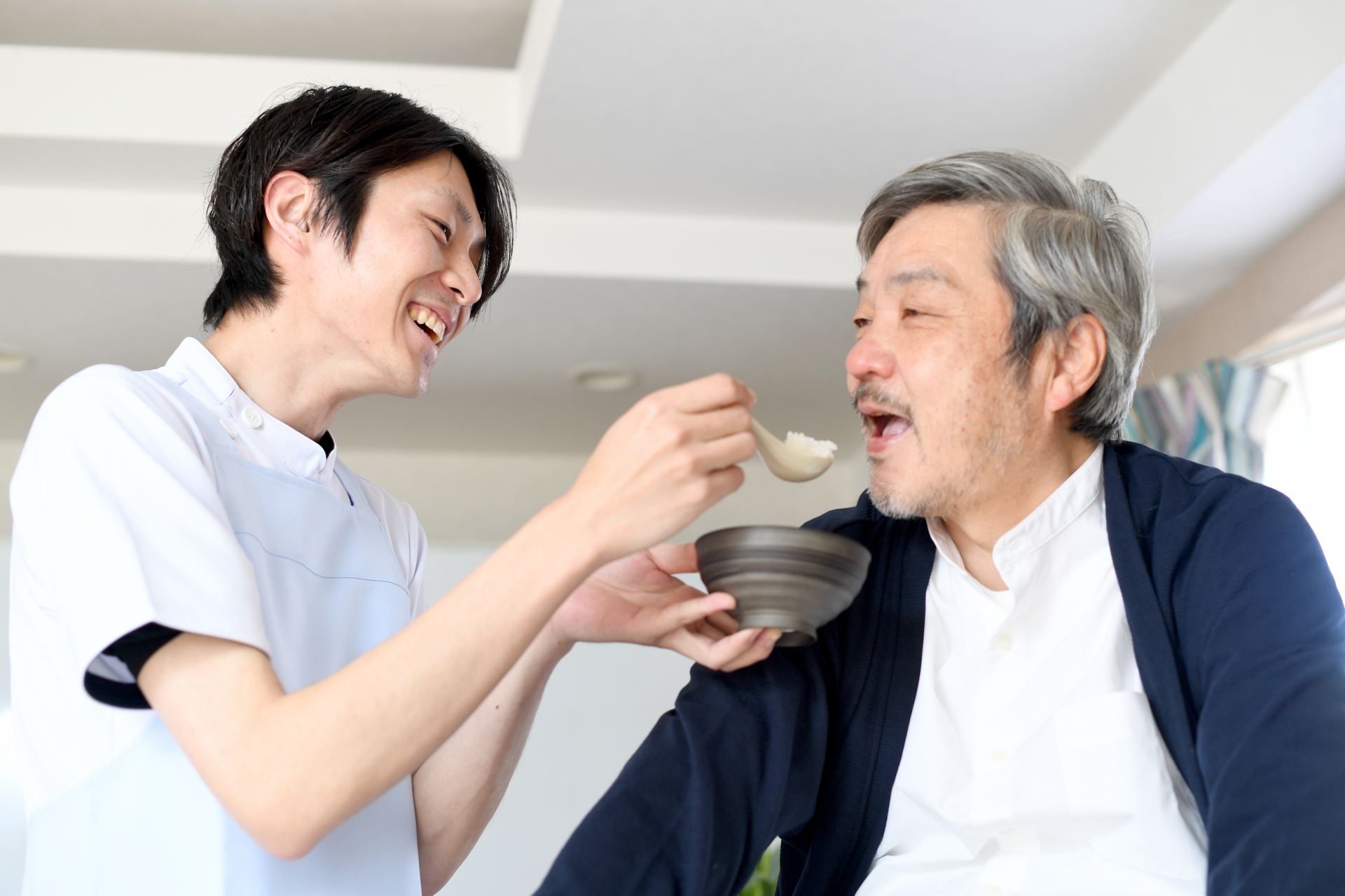 シニア男性に食事の介助をするエプロン姿の若い男性