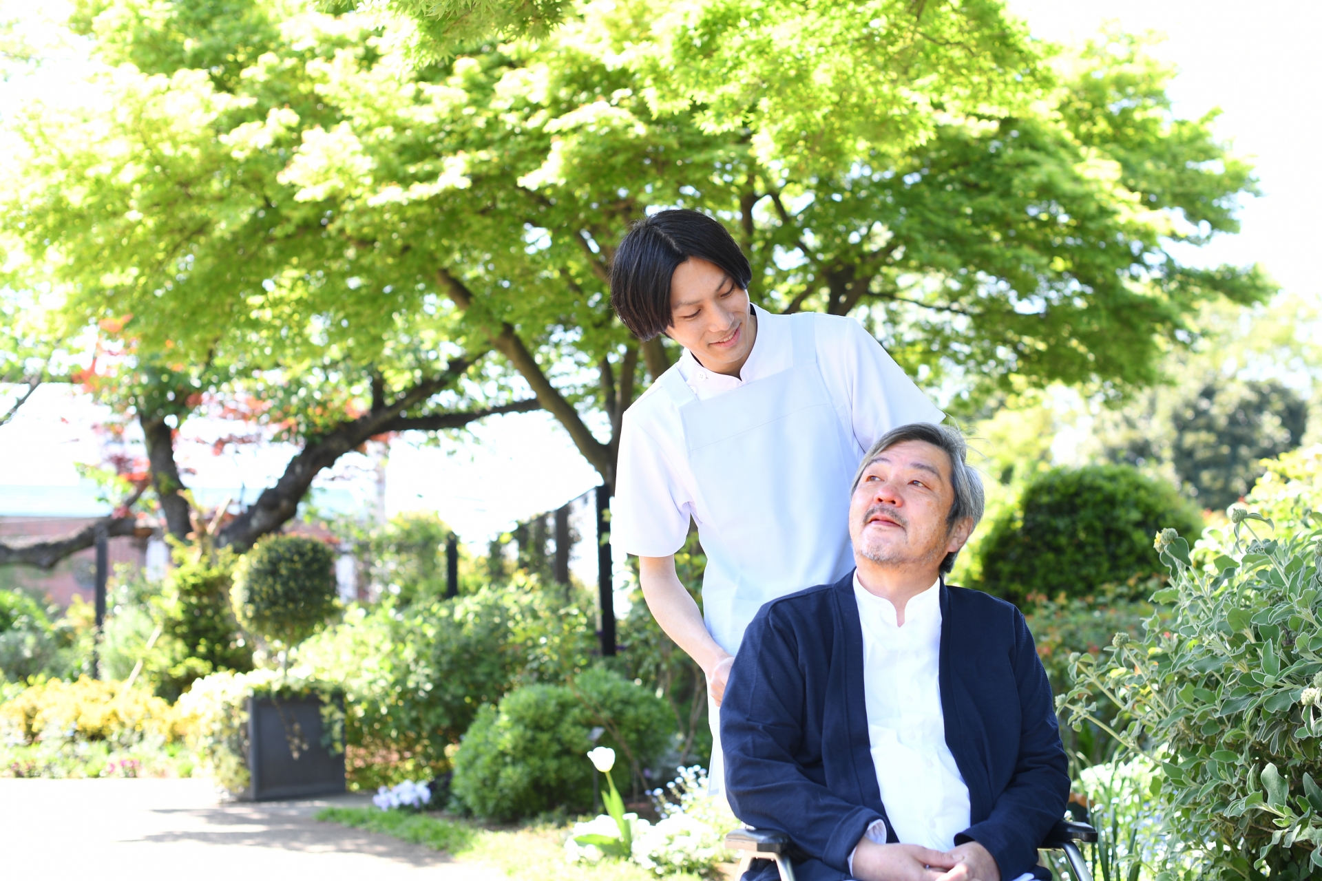 屋外で散歩をする介護ヘルパーの男性と車椅子のシニア男性