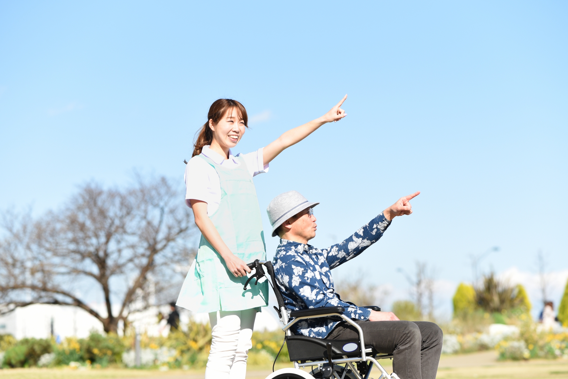 青空の屋外で車椅子を押す若い介護士の女性とシニア男性