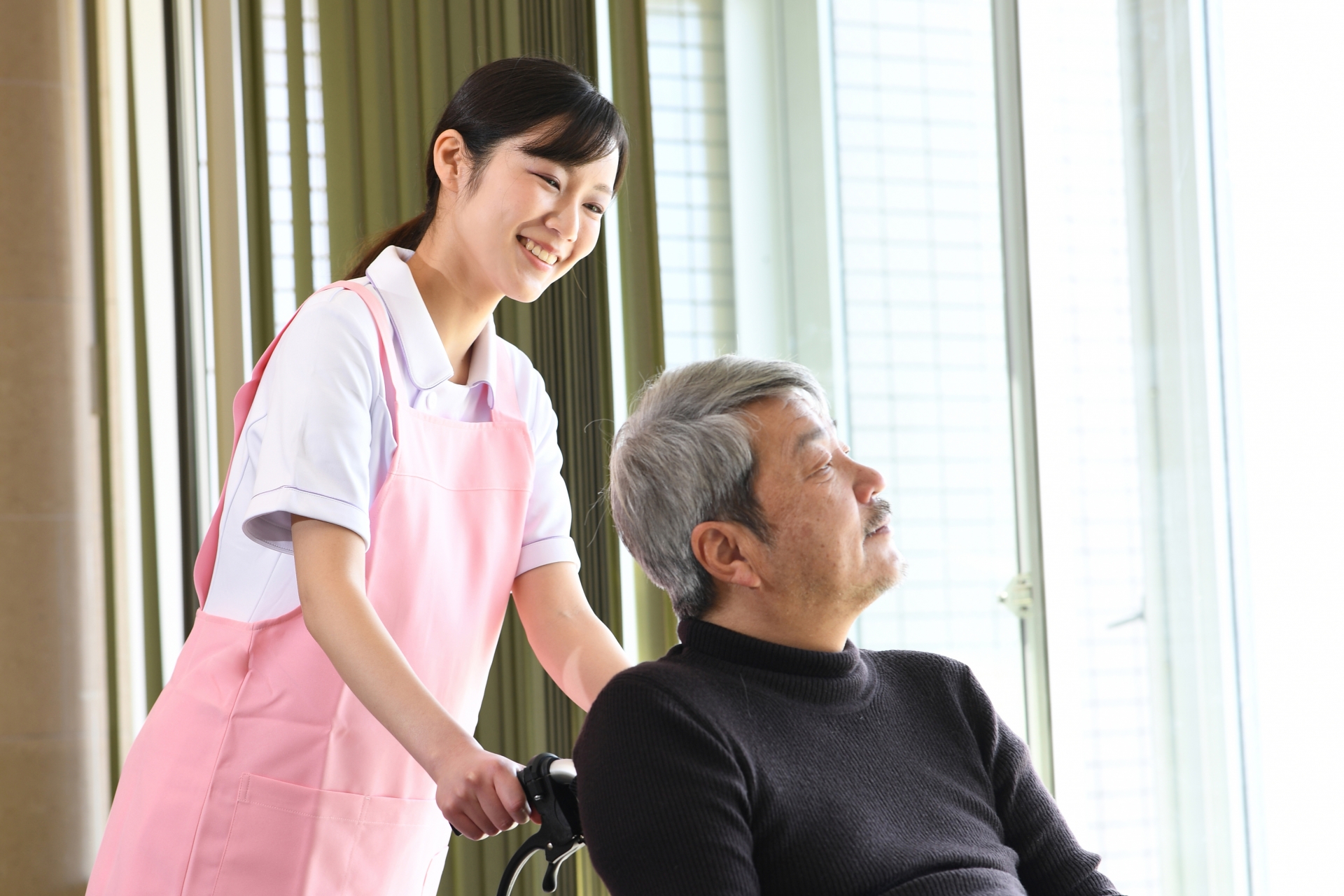 室内で車椅子を押す若い女性とシニア男性